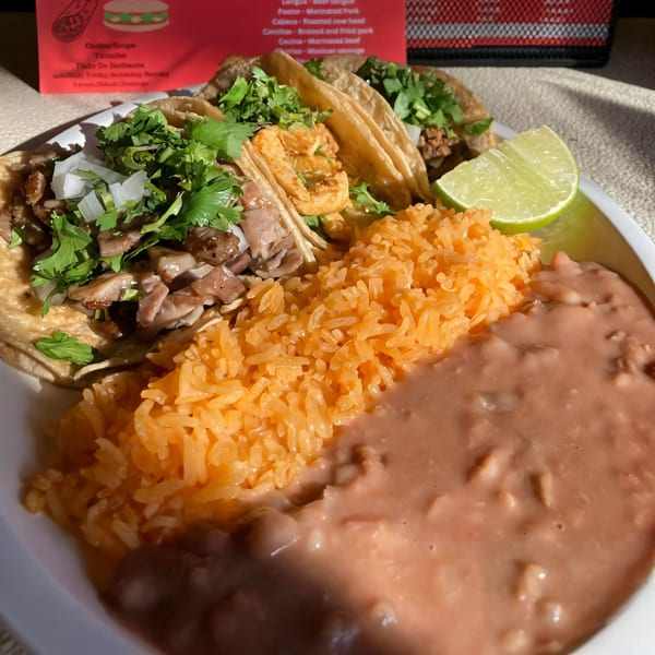 Three Mexican street tacos with cilantro, onions, rice, beans, and a lime wedge on a white plate.