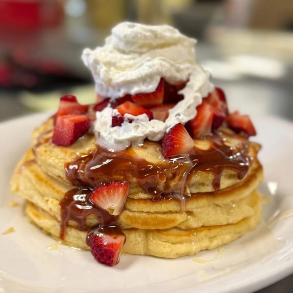 Stack of pancakes topped with whipped cream, strawberries, and caramel sauce on a white plate.