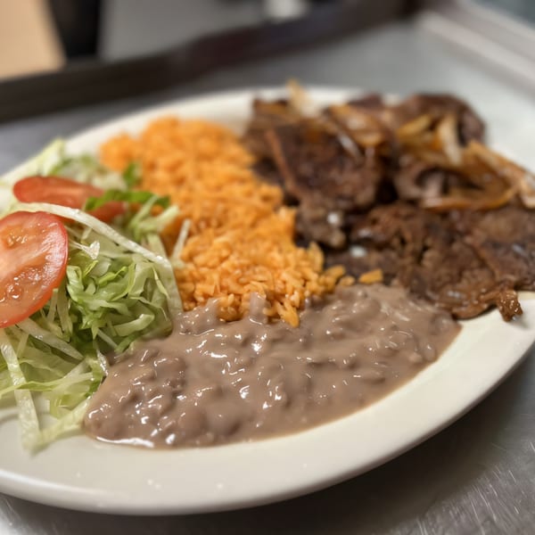 A Mexican dish with steak, rice, refried beans, shredded lettuce, and sliced tomatoes on a white plate.