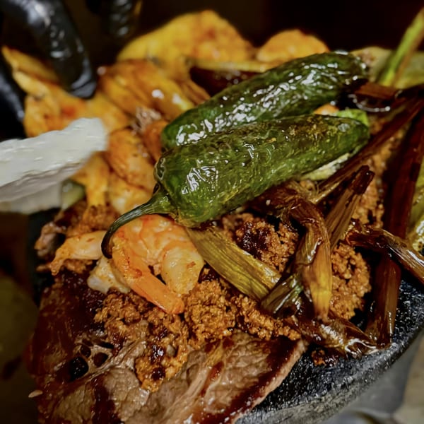 Close-up of a sizzling Mexican molcajete dish with steak, shrimp, chorizo, and green peppers.