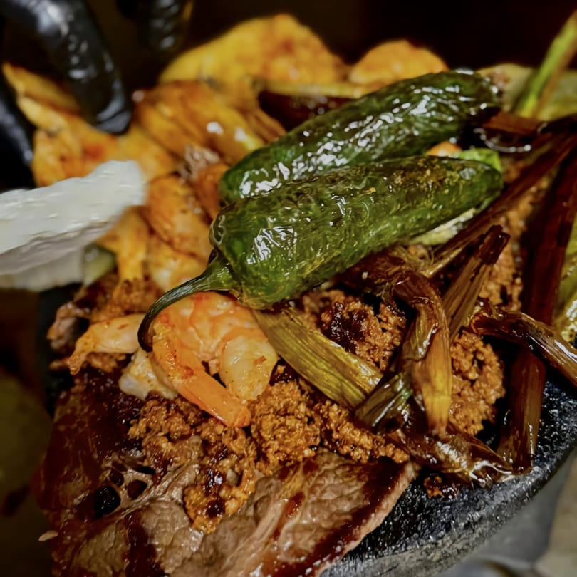Close-up of a sizzling Mexican molcajete dish with steak, shrimp, chorizo, and green peppers.