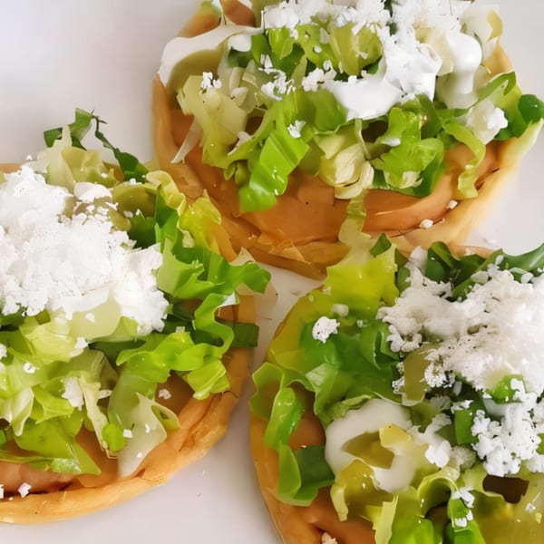 Three Mexican sopes topped with lettuce, crumbled white cheese, and crema on a white plate.
