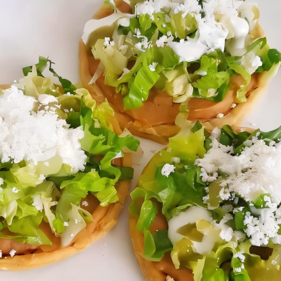 Three Mexican sopes topped with lettuce, crumbled white cheese, and crema on a white plate.
