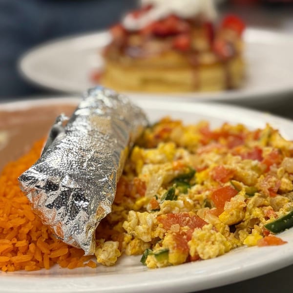 Mexican breakfast plate with scrambled eggs, rice, beans, and a foil-wrapped item, with pancakes in the background.