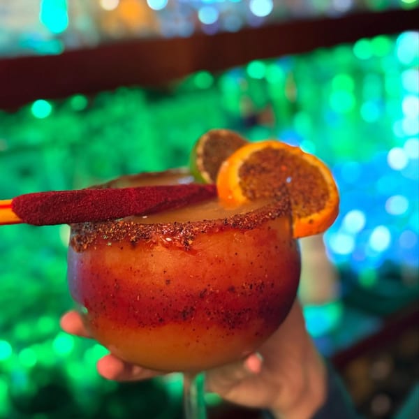 A hand holds a large, spiced Mexican drink garnished with orange and lime slices, against a blurred green background.