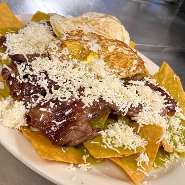 Chilaquiles with green sauce, steak, fried eggs, and shredded cheese on a white plate.